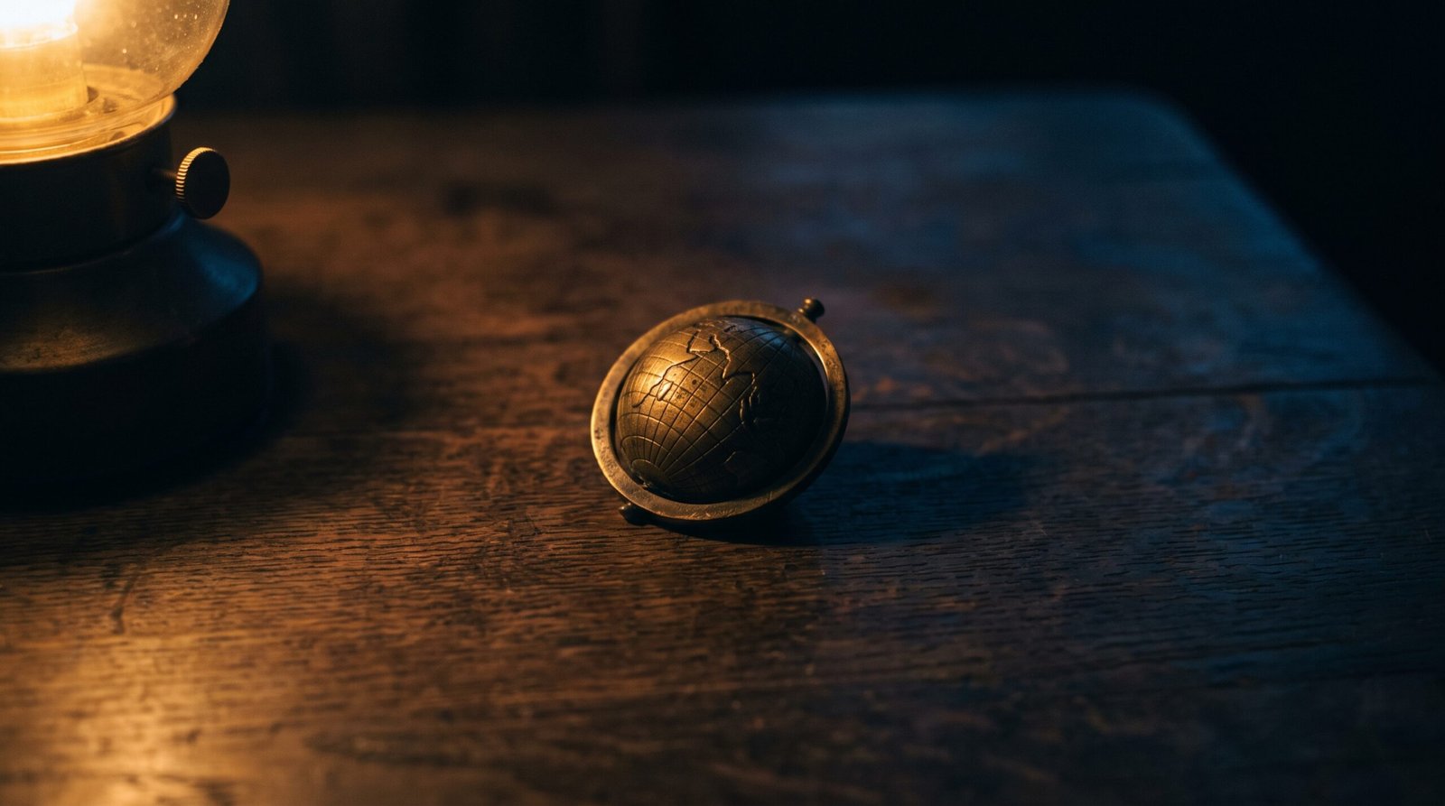 A single brass globe marker on a dark wood surface, dim warm amber side light, deep navy shadows, no people visible.